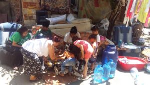 Women and children sorting belongings by water jugs